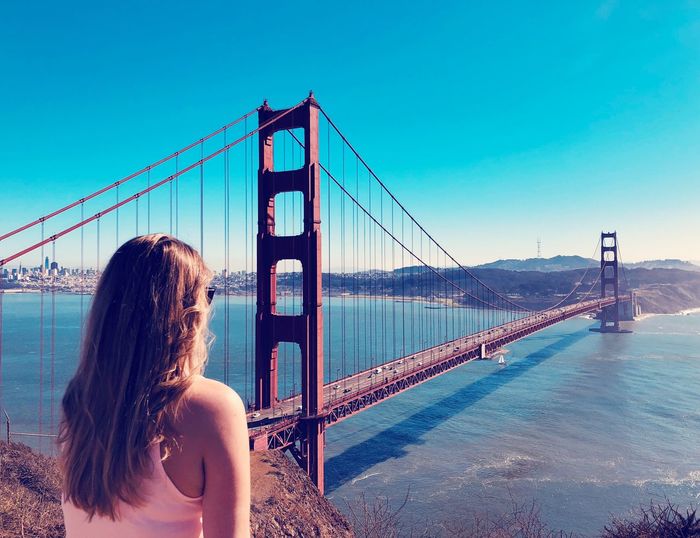 Rear view of young woman looking golden gate bridge over bay against clear blue sky
