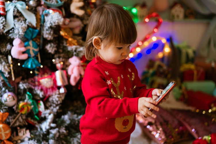 Little girl in red sweater is watching cartoons on phone next to christmas tree christmas tree
