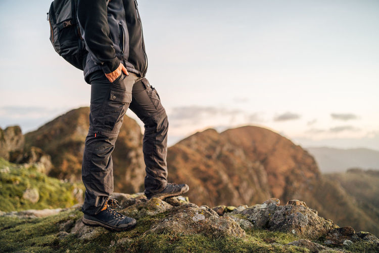 Side view of cropped unrecognizable lonely male explorer with backpack standing on top of green hill and contemplating nature while traveling through mountainous terrain