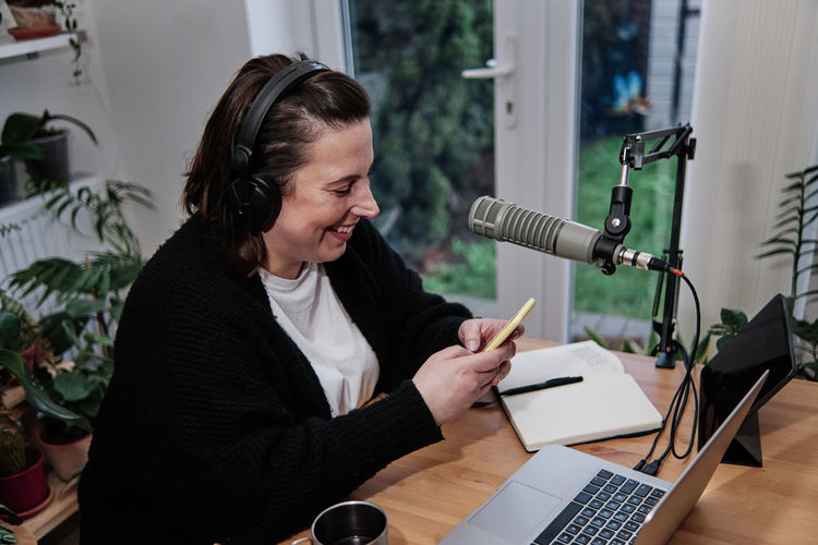 Smiling woman using smart phone by laptop at desk during podcast session at home