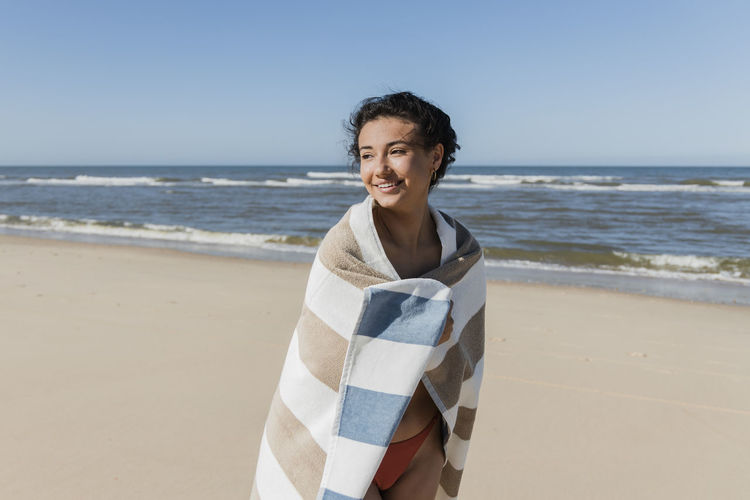 Smiling woman wrapped in towel looking away while standing at beach
