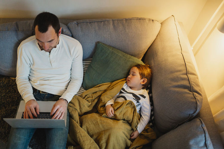 Father using laptop in living room while daughter sleeping next to him