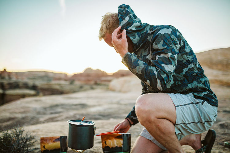 Male camper prepares camp dinner and pulls camo jacket over head