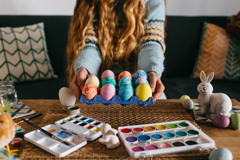 Unrecognizable female in sweater demonstrating carton with set of painted easter eggs while sitting on sofa