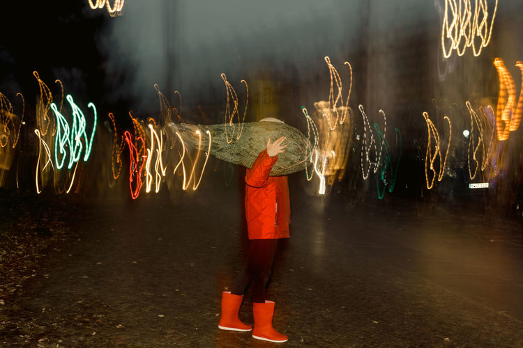 Girl carrying a christmas tree on her shoulders with street lights effect