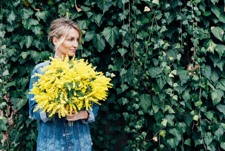 Stylish young woman in denim clothes with a bouquet of mimosa against the wall with bright foliage.