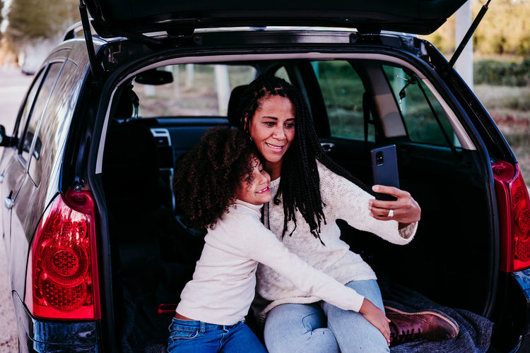 Smiling mother and daughter doing selfie while sitting at car trunk