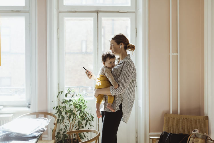 Businesswoman carrying baby boy while holding smart phone at home