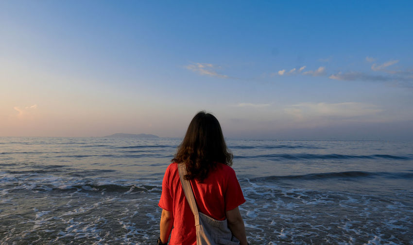 REAR VIEW OF WOMAN STANDING AT BEACH AGAINST SKY
