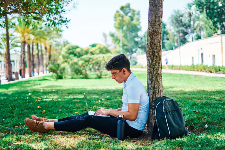 Side view of diligent male student doing homework while browsing netbook and sitting near tree on lawn in university campus